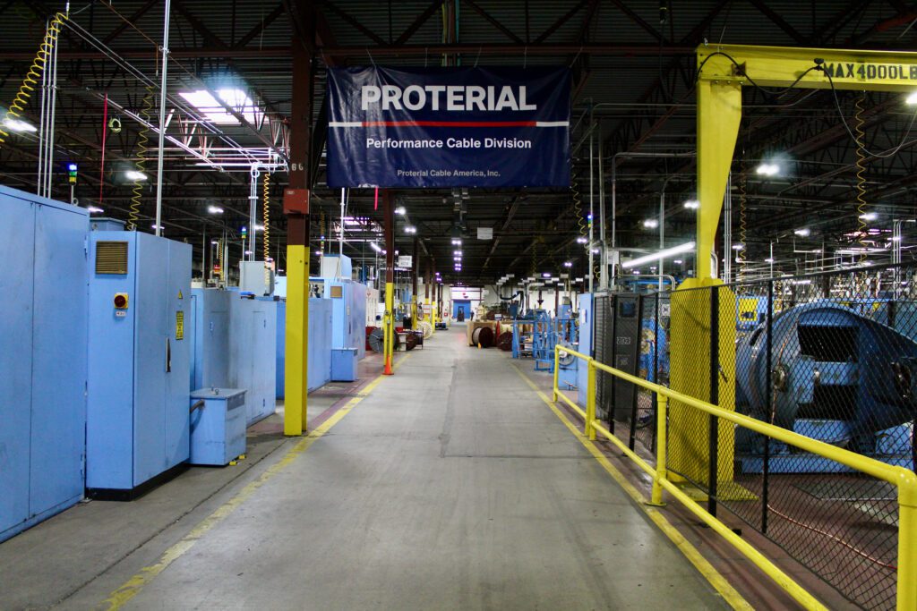 Proterial Banner hangs over pathway through factory floor with blue machines and yellow rails on either side.